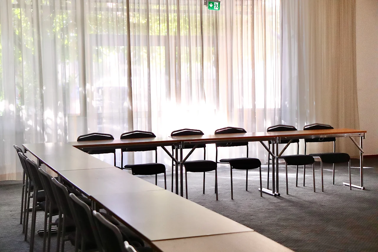 Long tables are arranged with chairs underneath in an empty conference room. Sunlight filters through sheer curtains, illuminating the carpeted floor.