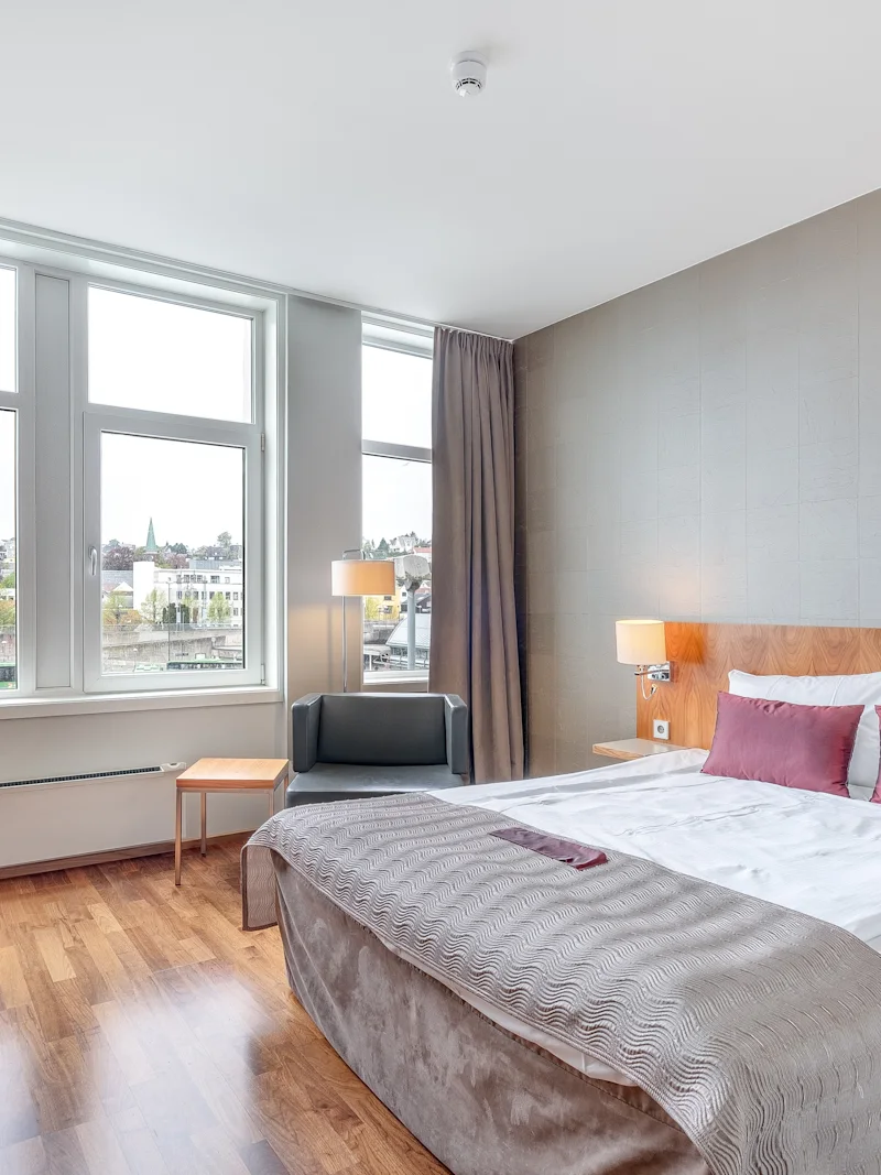 Katon mukaisesti pedattu hotellisänky, jossa on harmaa-valkoiset vuodevaatteet, sängynpöytien vieressä, sijaitsee suurten kaupunkinäkymää tarjoavien ikkunoiden vieressä, luoden valoisan ja modernin huoneilmapiirin.
