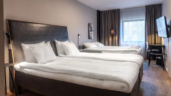 Two neatly made beds stand in a stylish hotel room, featuring dark curtains, wall-mounted TV, desk, and soft lighting enhancing a comfortable ambiance.