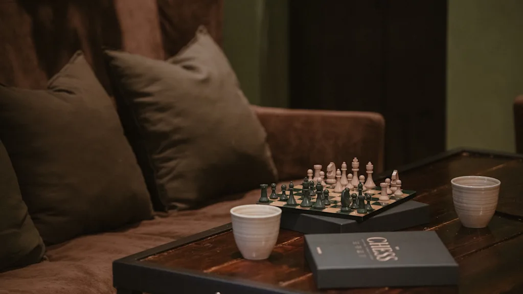 Viihtyisä näkymä, jossa shakkilauta on asetettu puupöydälle, kahden kupin ja CHESS-merkityn laatikon kera, mukavan ruskean sohvan vieressä.