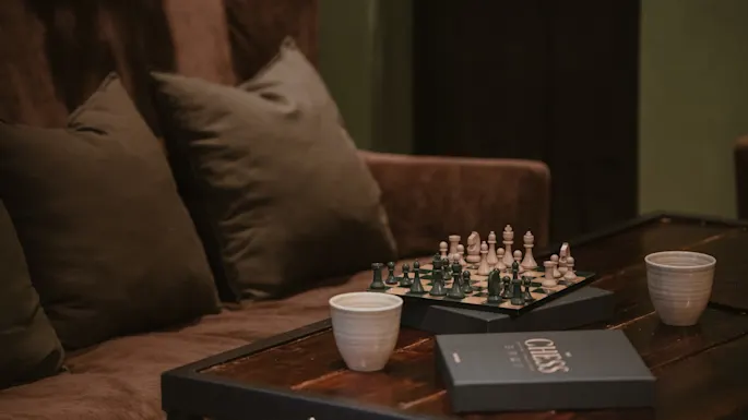 En koselig scene med et sjakkbrett satt opp på et trebord, sammen med to kopper og en eske merket CHESS, ved siden av en komfortabel brun sofa.