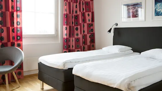 Two neatly made twin beds sit side by side in a bright room with red patterned curtains, white walls, and framed artwork. A modern chair is nearby.