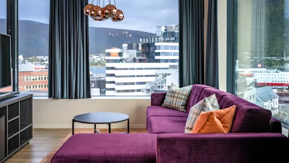 En stilig stue med en lilla seksjonssofa, moderne belysning og panoramautsikt over by og fjell.