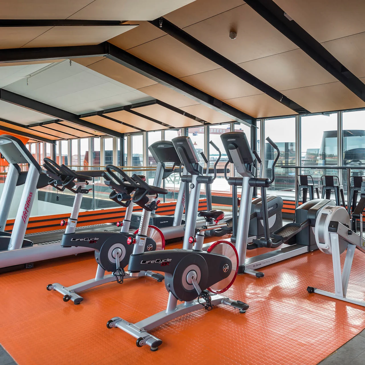 Treadmills and exercise bikes are placed on an orange floor mat in a modern gym with large windows showing an urban view.