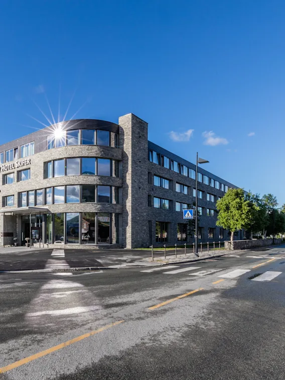 En modern, fler-våningsbyggnad märkt "Scandic Valdres Hotel" står i ett gatuhörn, med reflekterande fönster omgiven av träd, berg och en klarblå himmel.