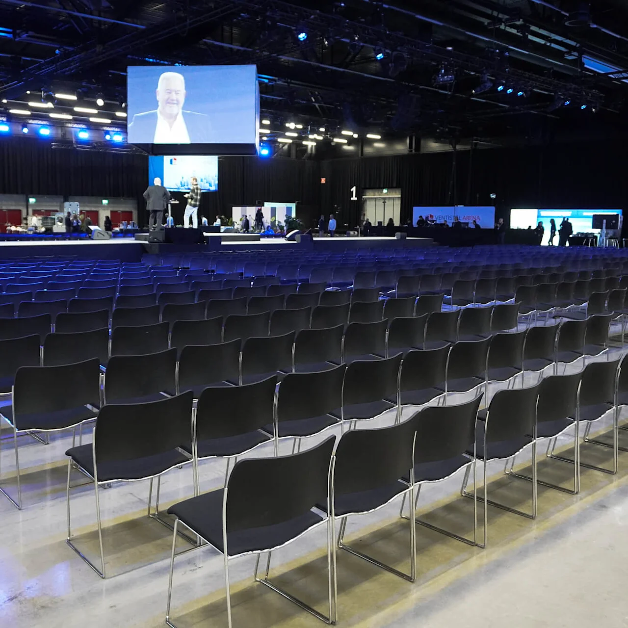 Rader av tomma stolar vetter mot en scen i en stor konferenshall med blå belysning. En skärm visar en talande person. Folk sätter upp utrustning i bakgrunden.