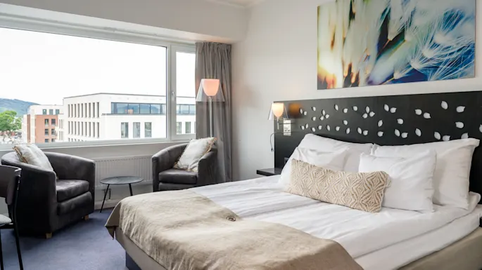 A neatly made bed with white linens and beige blanket; surrounded by armchairs and table near a large window, artwork above the headboard in a bright, modern hotel room.