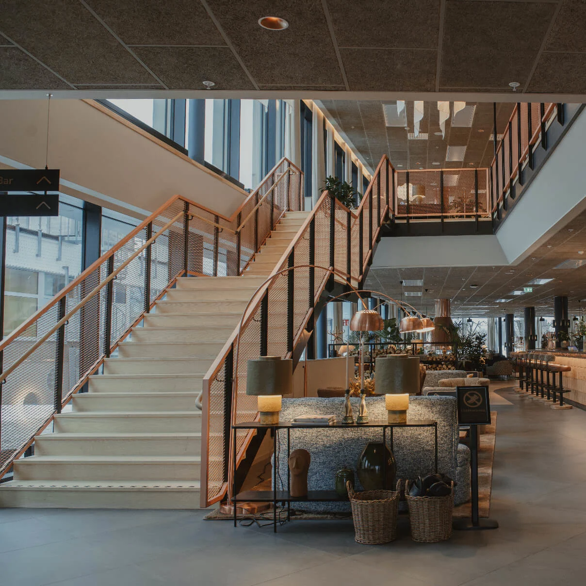 Stor trappa reser sig i en modern lobby, flankerad av en elegant bar, trähyllor och sittgrupp. Mjuk belysning och växter förstärker den inbjudande atmosfären.