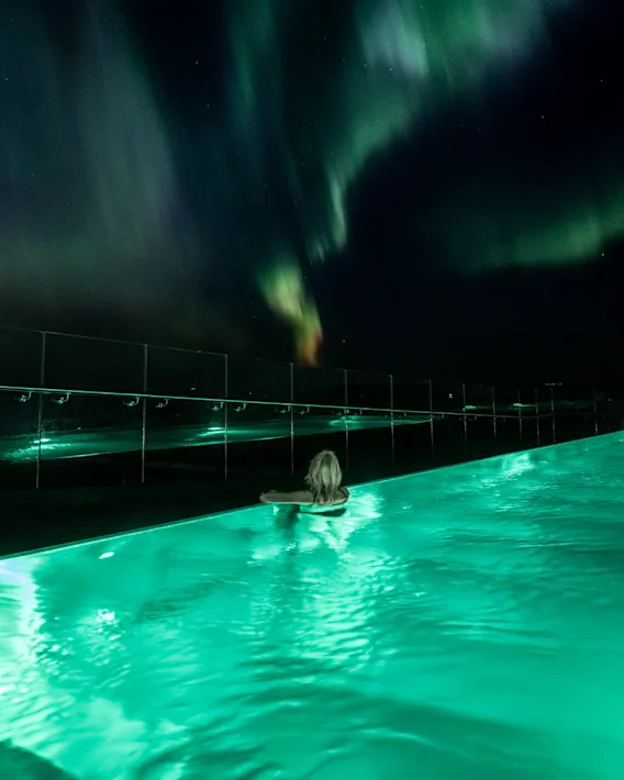 En person nyder en svømmetur i en glødende grøn pool om natten, med det levende nordlys dansende over den mørke himmel.
