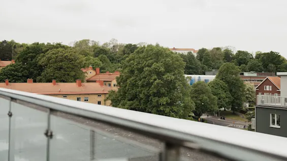 A glass railing overlooks a cityscape with red-roofed buildings and lush green trees under an overcast sky. The view suggests an elevated position, possibly a balcony.