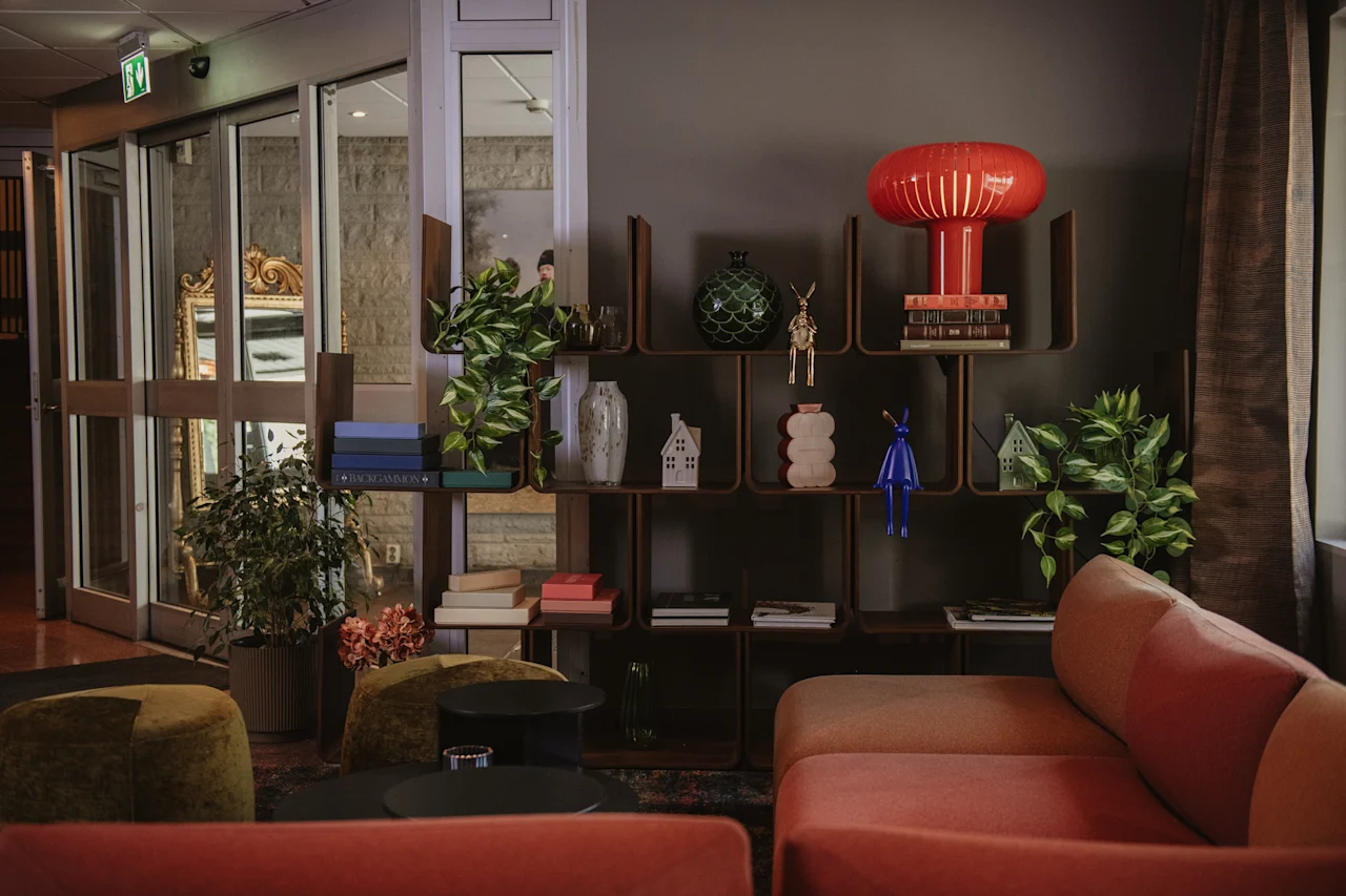 Et koselig salongområde med en dekorativ bokhylle, rød lampe og planter. Komfortable sitteplasser. VAGSAMMON, BACKGAMMON.