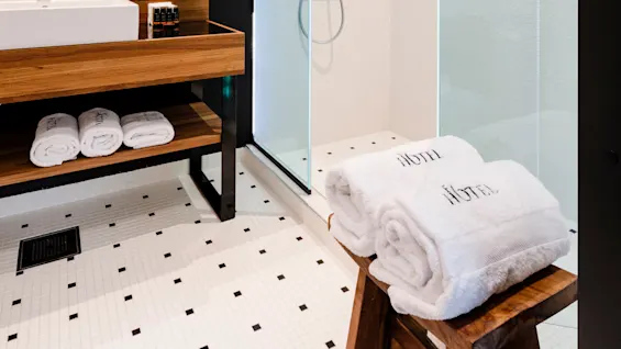 Et moderne bad har en glassinnelukket dusj, en trevask med en hvit vask og sammenbrettede håndklær merket "HOTEL". Sort-hvite armaturer kontrasterer i det moderne rommet.
