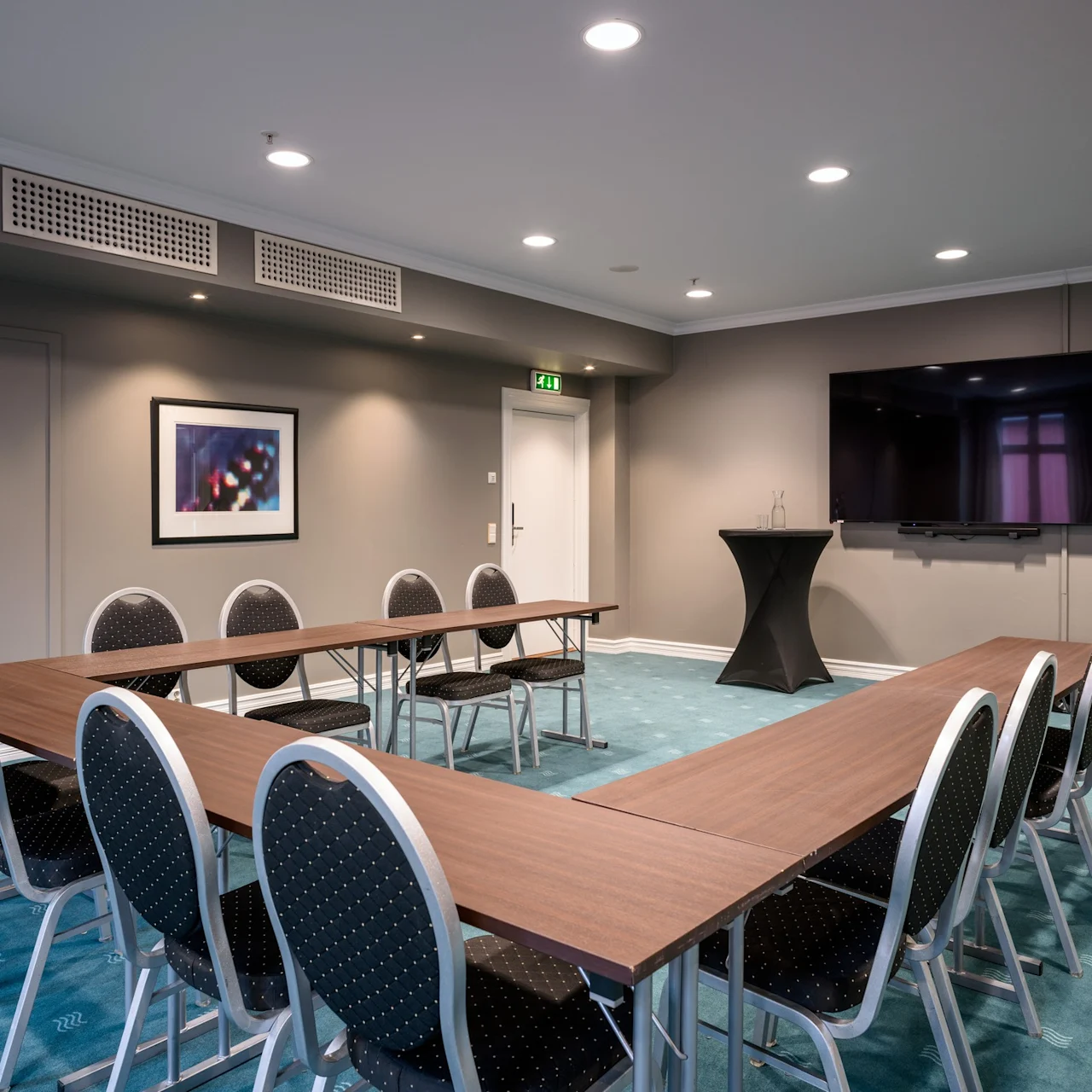 Bord arrangerade i U-form med stolar i ett konferensrum; en stor skärm och ett litet bord med två tomma flaskor finns i en mattsatt miljö.