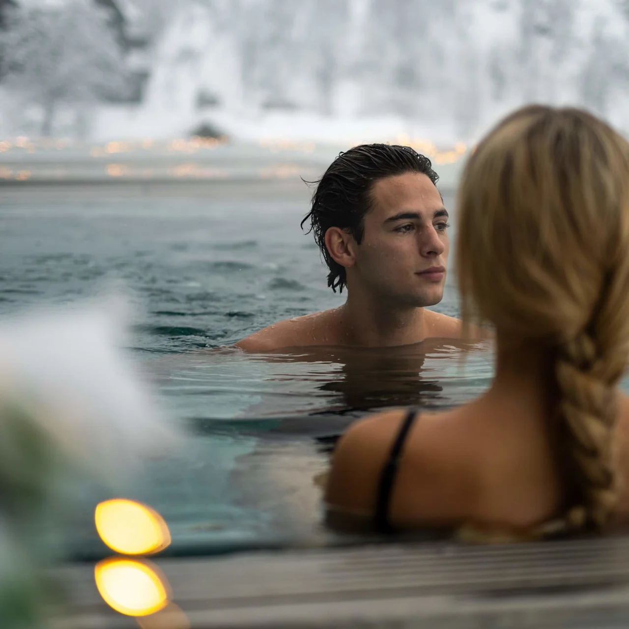 Man and woman relaxing in an outdoor hot spring, surrounded by snowy mountains. The woman's back is turned, her hair braided. The serene atmosphere features blurred fairy lights.