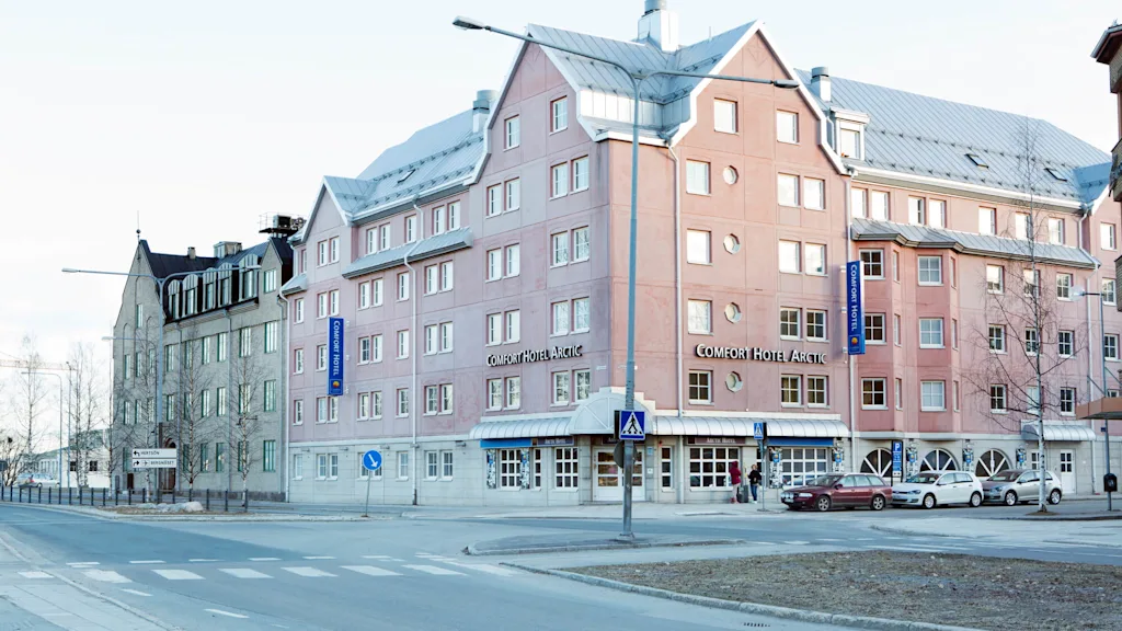 En laksefarvet hotelbygning med et klart blåt skilt, "Comfort Hotel Arctic," omgivet af en gade med parkerede biler og nøgne træer.