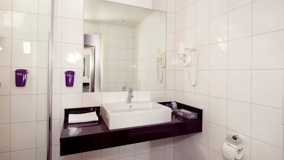 A modern bathroom with a white rectangular sink, mounted on a black countertop, and tiled walls, with two hairdryers and two purple soap dispensers.