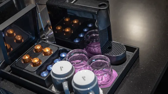 En Nespresso-kaffemaskine står på en mørk bordplade, omgivet af farverige kaffekapsler og klare glaskrus. Opsætningen er pæn og organiseret, klar til brug.