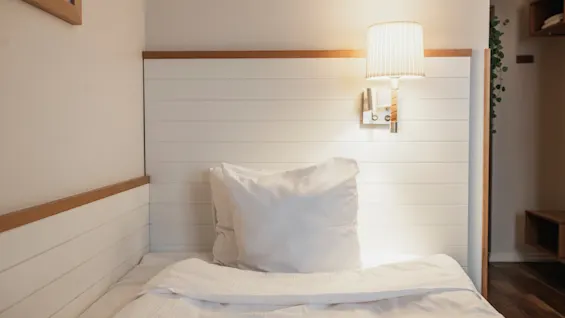 Ett mysigt sovrum med en prydligt bäddad säng, vita sängkläder och en vägglampa som ger varmt ljus.