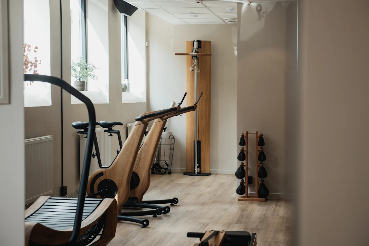 A bright fitness room at Home Hotel Uppsala with modern wooden exercise equipment, including a treadmill, bikes, and weights.