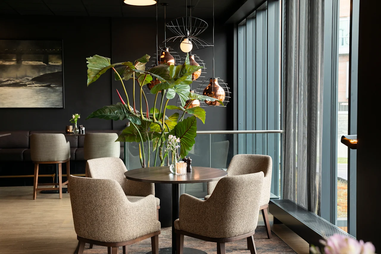 Et stilig restaurantinteriør med et rundt bord, komfortable stoler og en stor plante, satt mot en bakgrunn av mørke vegger og et vindu.