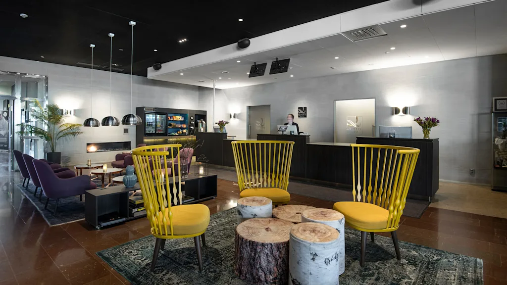 Reception med en person som arbejder, omgivet af stilfulde møbler i en moderne lobby. Gule stole og stubeborde komplementerer mørke sofaer, planter og hængelamper. Tekst: "QUALITY HOTEL."