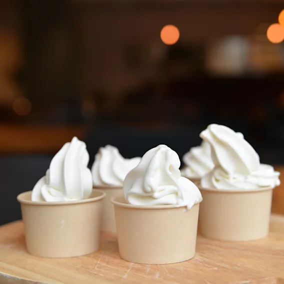 Four cups of soft-serve ice cream sit on a wooden surface in a warmly lit room with blurred background lights.