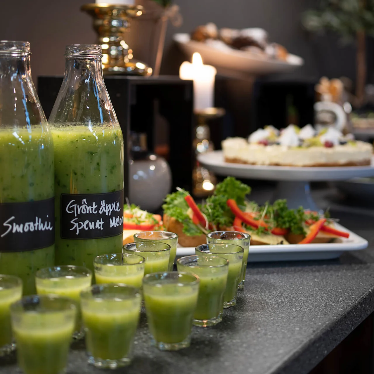 Flaskor med grön smoothie med texten "Smoothie" och "Grönt äpple, Spenat, Melon" på en bänkskiva, omgivna av ljus, snacks och en tårta i buffémiljö.