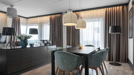 A dining table with green velvet chairs sits under modern pendant lights. Nearby, a sideboard holds a vase with flowers and drinking glasses. Neutral curtains frame large windows.
