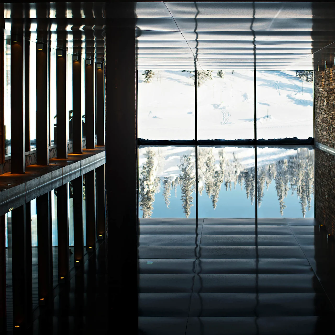 An indoor pool reflects a snowy landscape and trees, bordered by a stone wall and columns.
