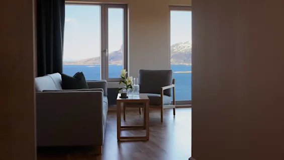 A gray sofa and chair sit around a small wooden table with flowers, facing large windows showcasing a sea view and distant mountains.