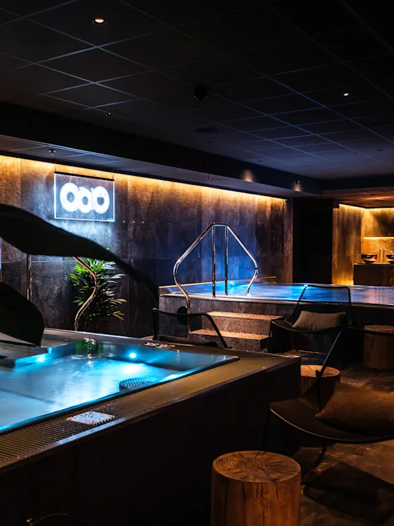 Luxurious indoor spa features illuminated pools, surrounded by ambient lighting and modern decor, including plants and seating. Neon sign with three circles on wall provides subtle illumination.