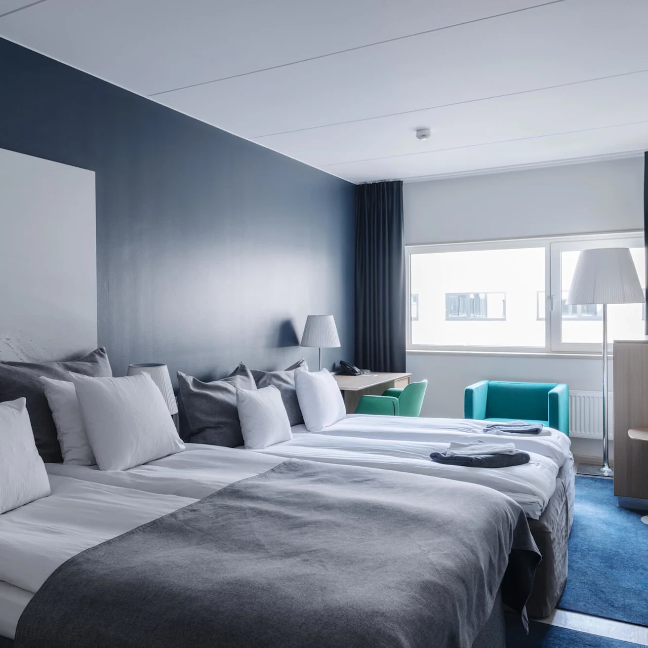 Ett modernt hotellrum med två prydligt bäddade sängar i grått och vitt, en stor landskapskonstverk ovanför, två turkosa stolar vid ett fönster och ett litet skrivbord med lampa.