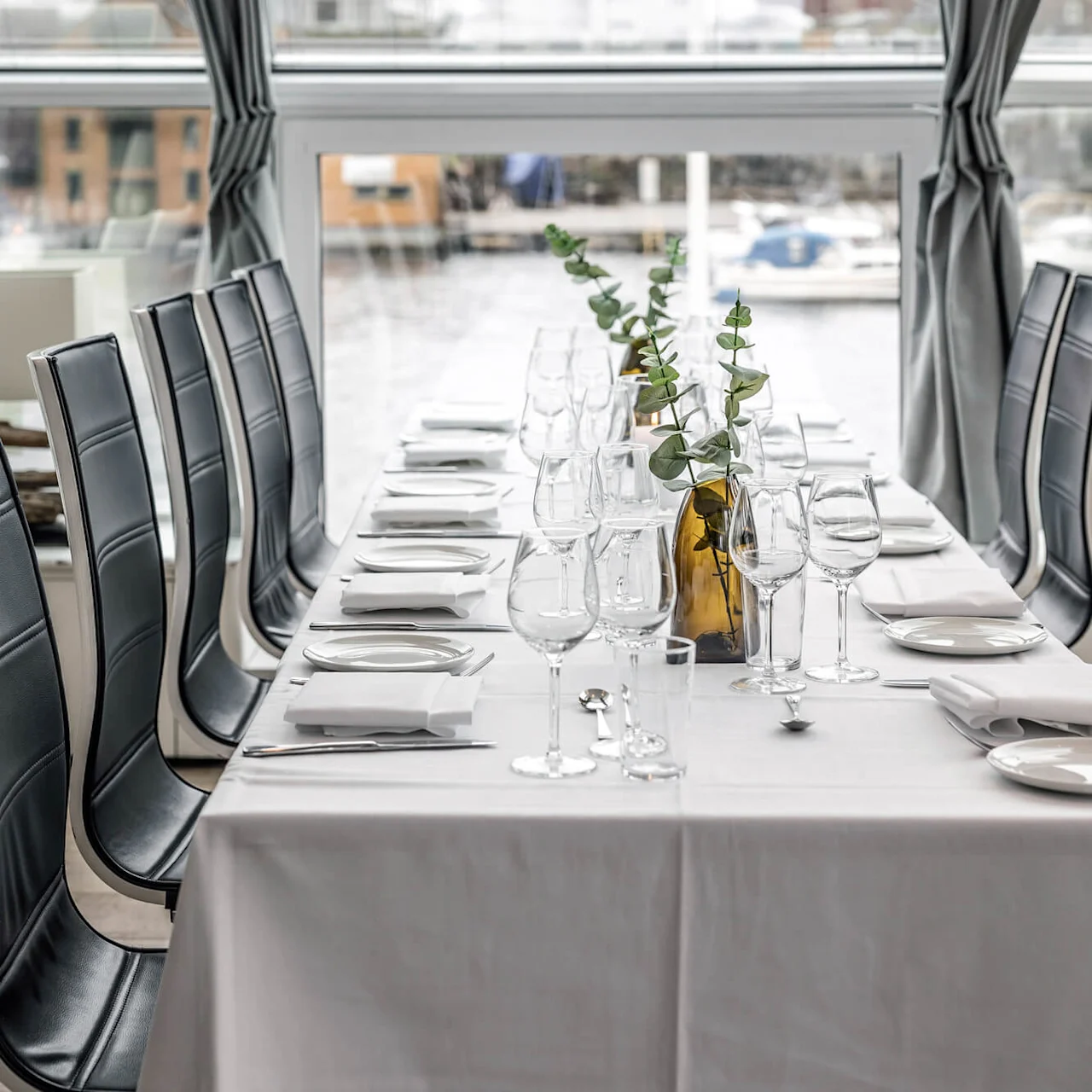 Elegant matbord med vita dukar, glas, tallrikar och bestick. Grönt i bärnstensfärgade vaser dekorerar mitten. Omgivet av svarta stolar, med fönsterutsikt över båtar utanför.