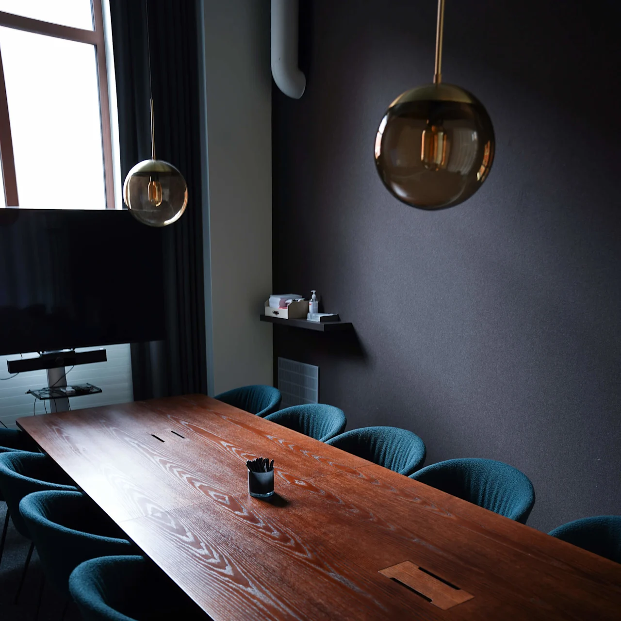 A long wooden conference table is surrounded by blue chairs beneath spherical pendant lights in a modern meeting room with a wall-mounted screen and a window.
