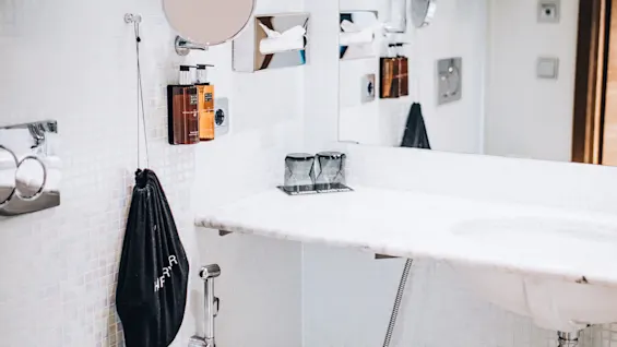 En badeværelsesvask med et marmorbassin, vægmonterede toiletartikler, et lille rundt spejl og en papir-essunser. To glas står på vasken. En sort hængende taske er mærket "HERR".