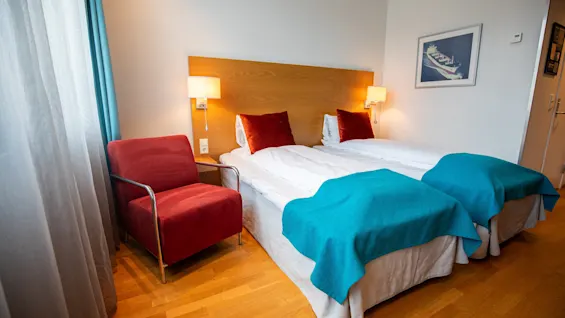 Et hotellrom med to senger har turkise tepper og røde puter, en rød lenestol ved et vindu med gardiner, tregulv og et innrammet bilde av et skip på veggen.