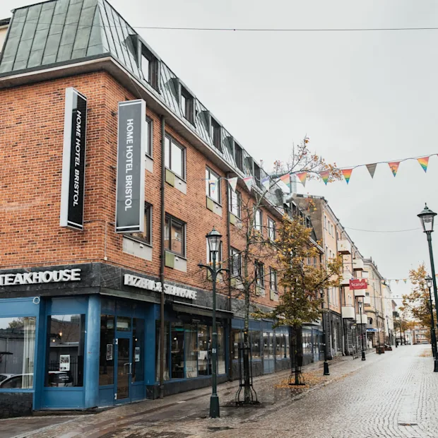 Katunäkymä tiilirakennuksella, jossa on hotelli ja ravintola. Värikkäät viirit roikkuvat märän mukulakivikadun yllä.
