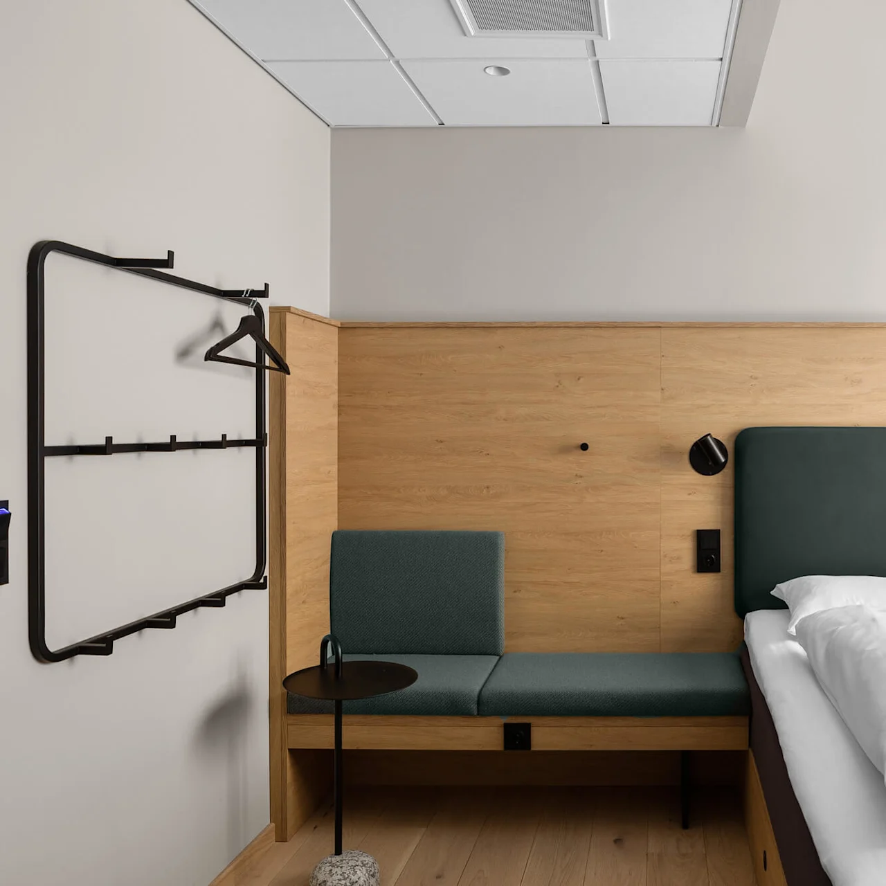 A neatly made bed with white linens sits beside a cushioned bench against a wooden headboard. A minimalist clothes rack and two pendant lights complete the modern, airy room.