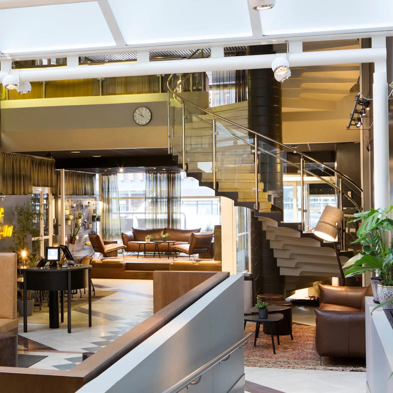 En elegant hotellobby med moderne møbler, et skilt for selvbetjening og en spiraltrappe, der fører op til et øvre niveau.