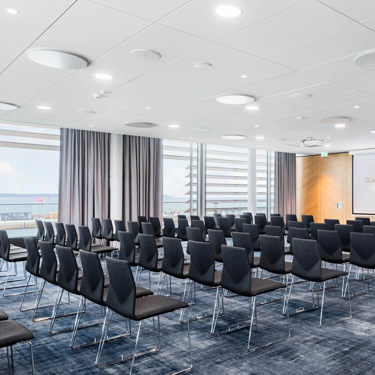 Svarte stoler vendt mot en projeksjonsskjerm med teksten "CLARION HOTEL SEA U" i et moderne konferanserom med store vinduer og grå gardiner. Skandinaviske flagg synlige utenfor.