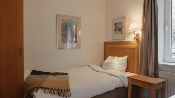 En hyggelig enkeltseng på et hotelværelse med en sengegavl i træ, et sengebord, et varmt tæppe og indrammet kunst på væggen.