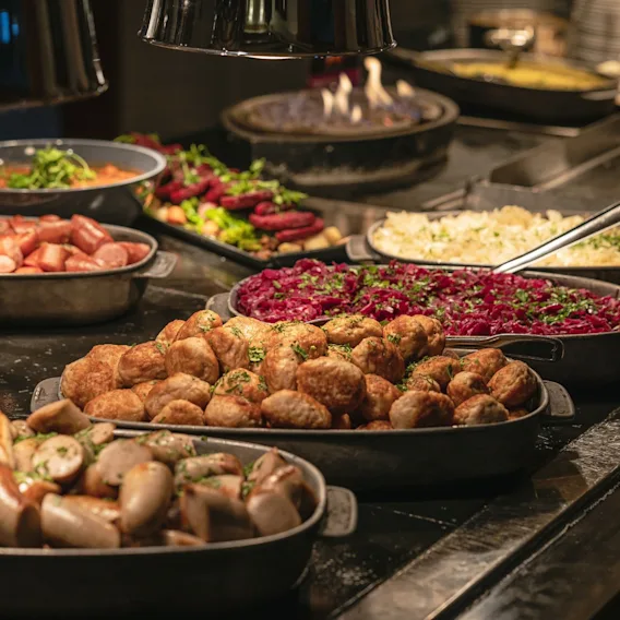 En julebuffet med fat fylt med kjøttkaker, pølser, rødkål og andre varme juleretter under varmelamper.