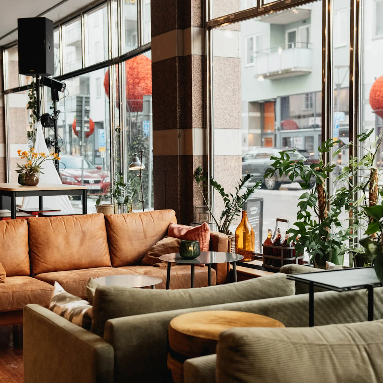Cozy couches in a cafe, next to large windows displaying a street view with cars. Potted plants decorate tables, and a stage with a red rope is visible. Signs outside read "ÖREBRO SALUHALL."