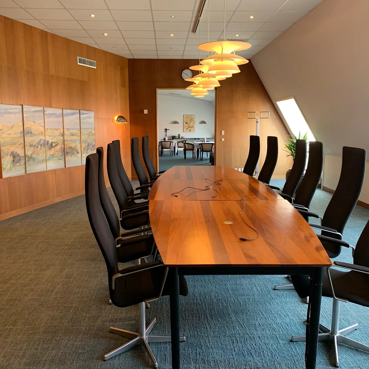 Long wooden conference table surrounded by black chairs, under hanging lamps, in a wood-paneled room with a painting and natural light from slanted windows.