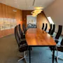 Long wooden conference table surrounded by black chairs, under hanging lamps, in a wood-paneled room with a painting and natural light from slanted windows.