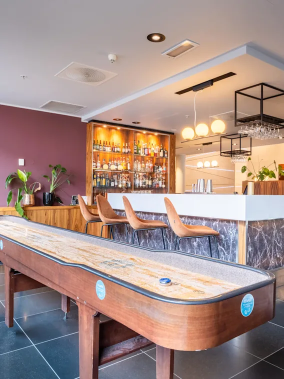 Bar med et shuffleboardbord foran, barstoler og en spritdisplay i en moderne lounge. Koselig belysning skaper en varm atmosfære.