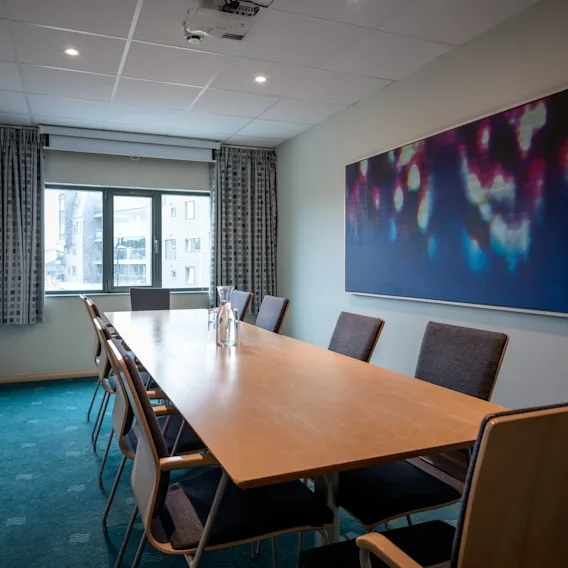Et langt trebord med åtte stoler er satt opp i et møterom. Scenen inkluderer en projektor i taket, et stort abstrakt maleri og et vindu med gardiner.