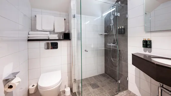 A modern bathroom features a glass-enclosed shower, white towels on shelves, and a sleek toilet against white tile. Two dispensers on the wall are labeled "hair & body."