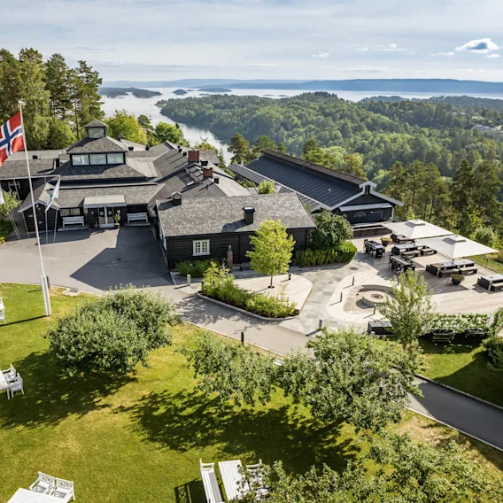 En stor bygning med norske flagg veier i et frodig, grønt landskap med utsikt over et naturskjønt vann. Hvite benker og trær pryder gresset foran.