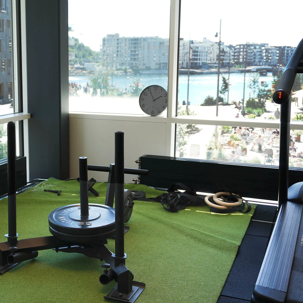A weighted sled sits on artificial grass indoors, with large windows showing a cityscape and water outside. A wall clock reads 1:15.
